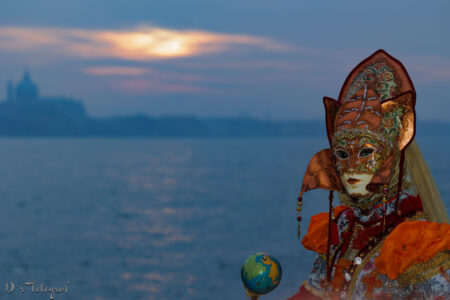 Eine Person in einem aufwendig verzierten venezianischen Karnevalskostüm mit einer kunstvollen Maske steht vor einer Wasserlandschaft bei Sonnenuntergang. Die Maske ist mit goldenen und roten Details verziert, und das Kostüm hat auffällige orangefarbene und rote Elemente. In der Hand hält die Person einen kleinen Globus. Im Hintergrund ist eine verschwommene Silhouette einer Kuppel und eines Gebäudes in Venedig, zu sehen.