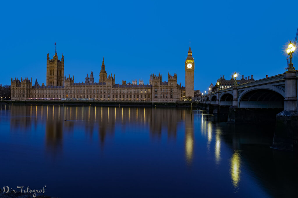 Houses of Parliament und BigBen in der Blauen Stunde am Morgen vom anderen Themseufer aus. Lichter spiegeln sich in der Themse.