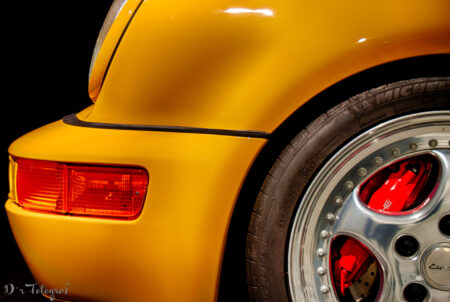 Detailaufnahme eines gelben Porsche 911 - von der Seite - Stoßstange und Teil des Rades sichtbar. Blinker in Orange und Bremssattel in rot stechen hervor.