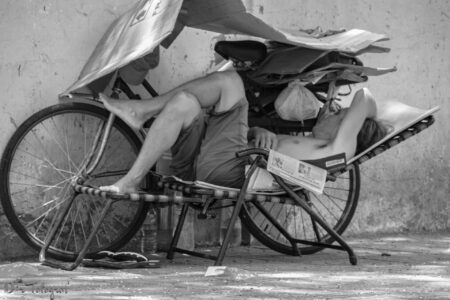 Mann schläft auf einer Klappliege vor seinem Fahrrad, welches mit Pappe abgedeckt ist, um einen Sonnenschutz für den Mann zu bieten. Schwarz-Weiss Bild