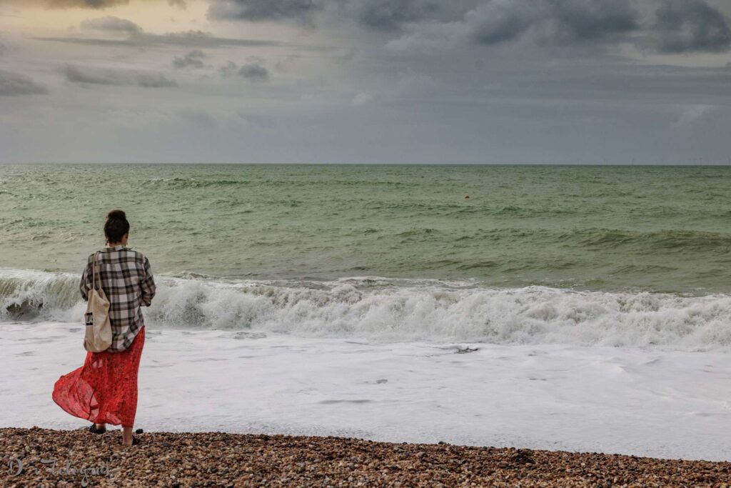 Frau mit rotem langen Rock steht an der Küste des Meeres und schaut auf das Meer. Der Himmel ist wolkenverhangen und ein wenig sieht man das Sonnenlicht durch