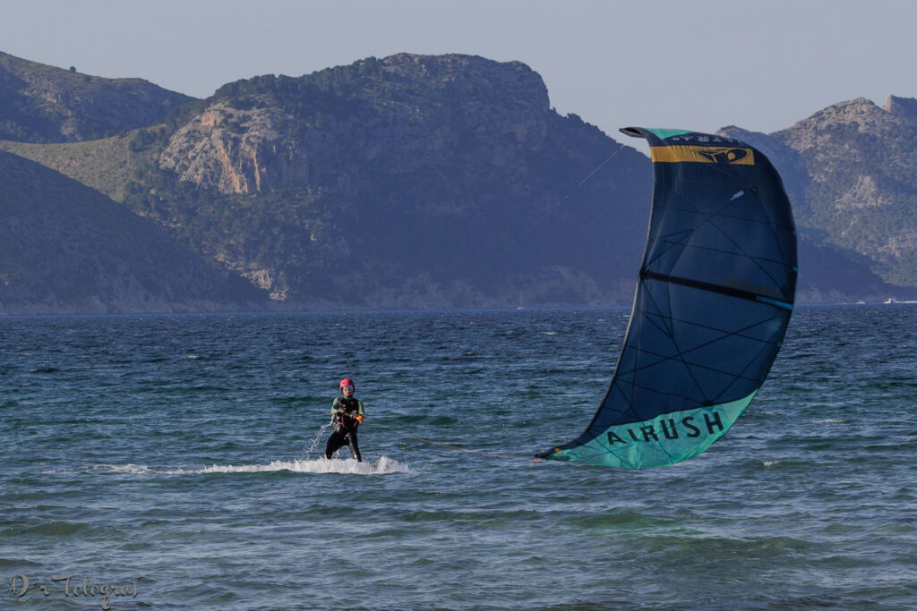 Das Bild zeigt eine Person, die Kitesurfen betreibt. Im Vordergrund ist der Surfer zu sehen, der auf dem Wasser steht und von einem Kite gezogen wird. Im Hintergrund erstreckt sich eine Berglandschaft über das Meer. Der Himmel ist klar und hell. Das Bild vermittelt ein Gefühl von Freiheit und Abenteuer.