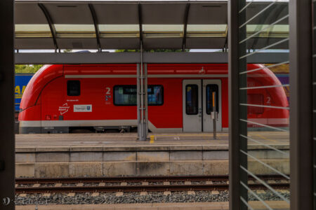 Roter Triebwagen der DB am Bahnsteig. Der Zug ist nicht ganz sichtbar, sondern nur ein kleiner Teil. Der andere wird von einer Glaswand verdeckt. In dieser spiegelt sich der sichtbare Teil des Zuges und erweckt so den Eindruck eines sehr kurzen Triebwagens.