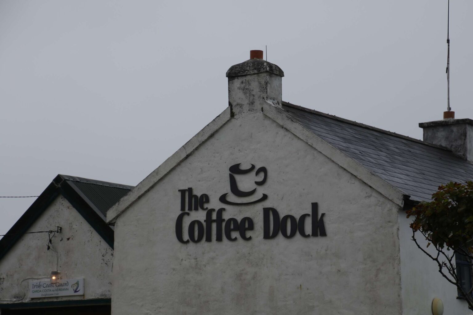Das Bild zeigt zwei Häusergiebel in hellgrau vor hellgrauem Himmel (Regenstimmung). Das vordere Haus hat am Giebel ein grosses Logo "The Coffee Dock" mit einer stilisierten Kaffetasse. Beim hinteren Haus, welches ein wenig versetzt und der Giebel nicht ganz sichtbar ist, brennt ein schwaches Aussenlicht über einem Schild mit der Aufschrift "Irish Coast Guard - Garda Costa na heireann-Knightstown Unit