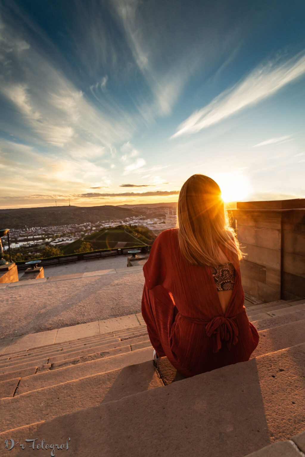 Eine blonde Frau sitzt auf einer Treppe, dem Betrachter den Rücken zugewandt. Der Blick geht über eine Stadt, der Himmel ist blau und hat Wolkenschleier. Recht neben dem blonden, langen Haar der Frau geht die Sonne unter