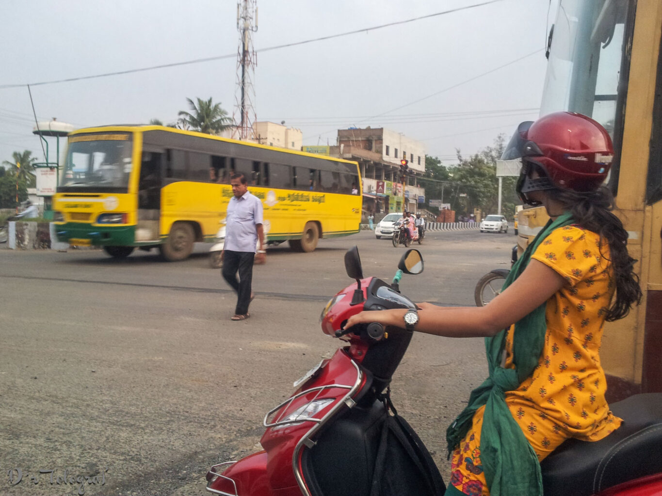 Bild zeigt eine Kreuzung in Chennai. Im Vordergrund wartet eine Frau in gelbem Kleid und dunkelrotem Motorradhelm auf einem dunkelroten Motorroller neben einem Bus darauf, dass sie losfahren kann. Ein Mann läuft mitten über die Kreuzung während im Hintergrund ein gelber Bus hinter dem Mann abbiegt.