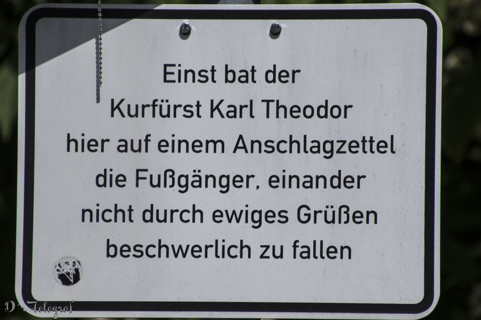 Bild zeigt ein weißes Blechschild mit schwarzer Schrift: »Einst bat der Kurfürst Karl Theodor hier auf einem Anschlagzettel die Fußgänger, einander nicht durch ewiges Grüßen beschwerlich zu fallen.«