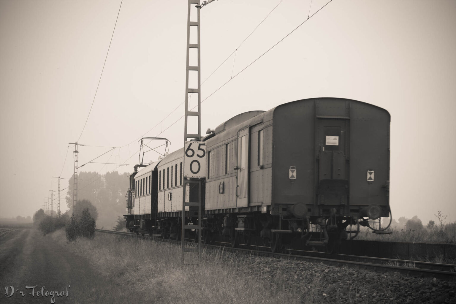 Sepia-Schwarzweiß-Aufnahme zeigt einen alten Zug, der auf Gleisen in einer nebligen Landschaft fährt. Der Zug besteht aus einer Lokomotive und zwei Waggons. Ein Mast der Oberleitung trägt ein Schild mit der Zahl „65 0“. Im Hintergrund sind Bäume und eine neblige Landschaft zu sehen.