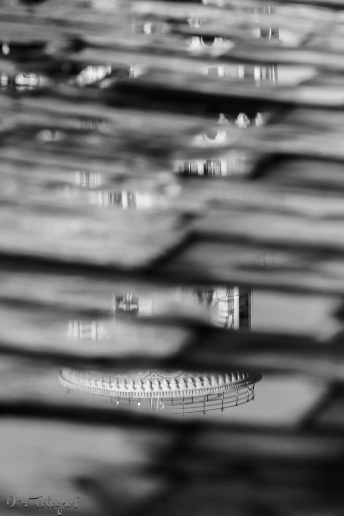 Schwarz-Weiß-Aufnahme. Blick auf mehrere Pfützen, die in einer Anordnung von Steinplatten liegen. Die Pfützen spiegeln verschiedene Strukturen des Turm von Pisa wider, wobei die deutlichste Spiegelung eine kuppelförmige Struktur mit einem Geländer und einem Turm im Hintergrund zeigt.