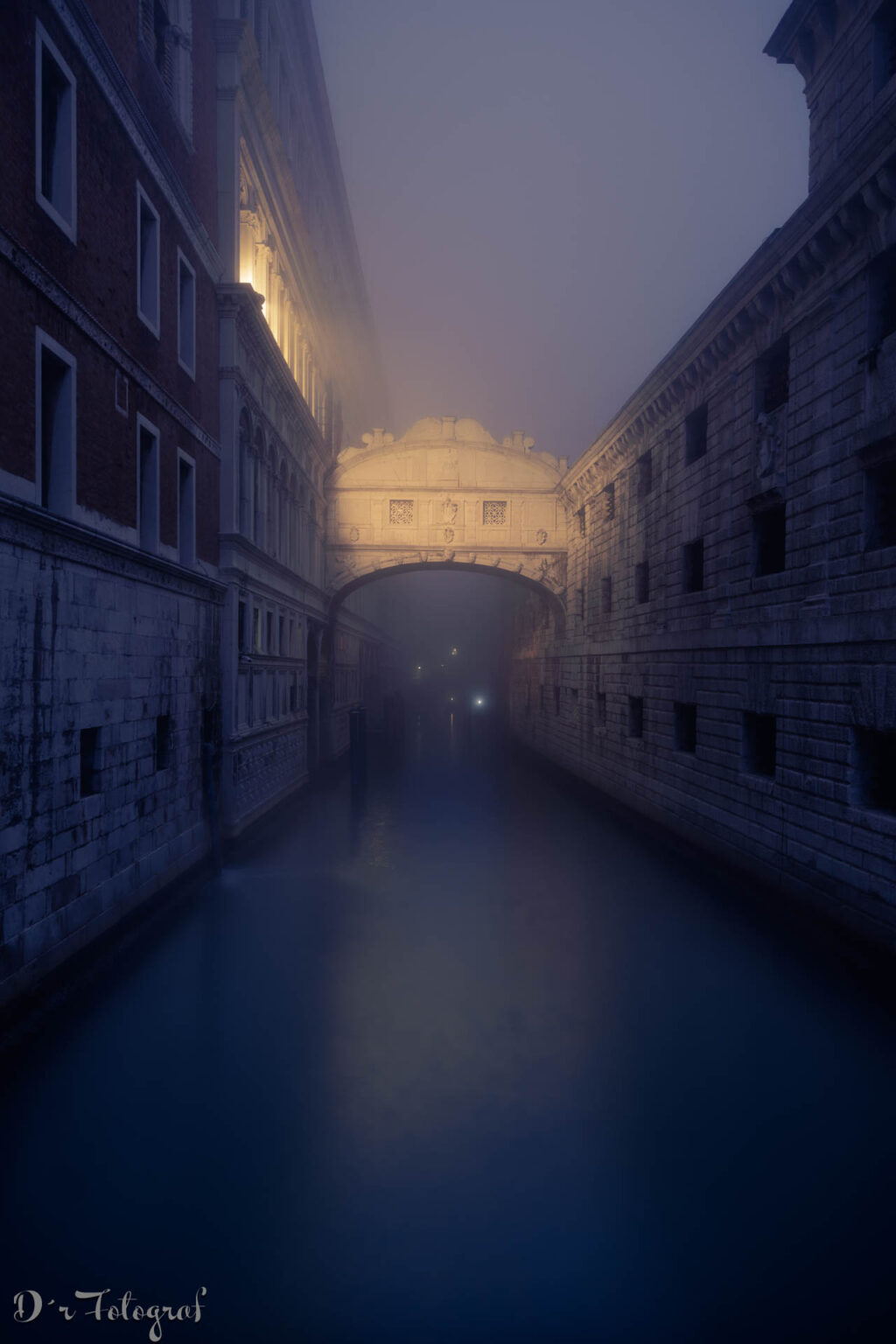 Ein vertikales Foto bei Nacht, das einen nebligen Blick den Rio di Palazzo Kanal hinunter auf die Seufzerbrücke in Venedig zeigt. Der Kanal füllt den größten Teil des unteren Vordergrunds und spiegelt das düstere, blaue Umgebungslicht der Nacht und des Nebels wider. Auf beiden Seiten des Kanals erheben sich hohe Gebäude. Links ist ein dunkles, rötliches Gebäude zu sehen, bei dem im Obergeschoss warme Lichter brennen und schwach auf den Nebel scheinen. Rechts ist ein helleres Gebäude mit einer Steinfassade und mehreren kleinen Fenstern zu sehen. Am Ende des Kanals ist die Seufzerbrücke zu sehen, eine geschwungene Brücke mit einer Steinfassade und einer kleinen Kuppel, die den Kanal überquert und in einem nebligen, helleren Licht erstrahlt. Die Brücke ist auch durch warme Lichter von innen beleuchtet, was einen Kontrast zum dunklen, nebligen Umgebungslicht schafft. Unter der Brücke ist eine weitere Brücke zu sehen, die in einem hellen, nebligen Licht erstrahlt. Der Nebel verleiht dem Bild eine mystische und geheimnisvolle Atmosphäre.