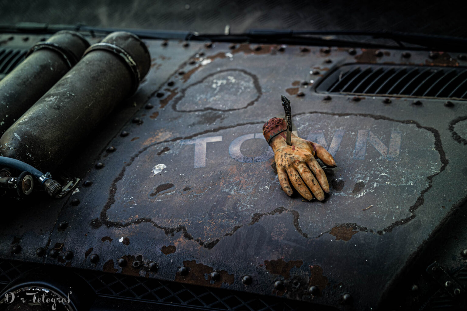 A close-up of a weathered, rusty hood from an post apocalypse vehicle. Two old, cylindrical tanks with hoses are mounted on the left edge. The word “TOWN” is visible on the metal in faded white letters. An artificial, dirty prosthetic hand is attached to the surface with a knife, positioned directly above the word. The metal plates are roughly riveted and show heavy signs of wear.